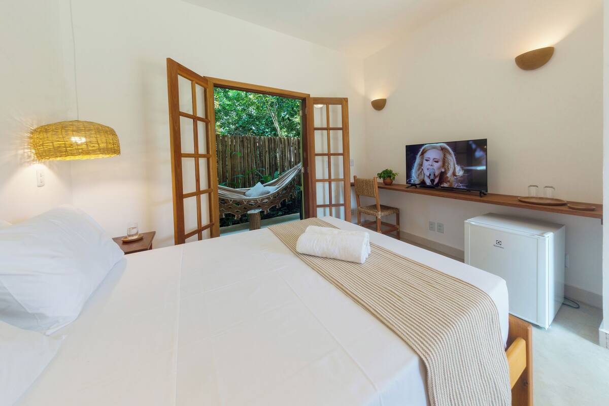 A bedroom with a bed, a television, a small refrigerator, and a view of greenery through an open door.