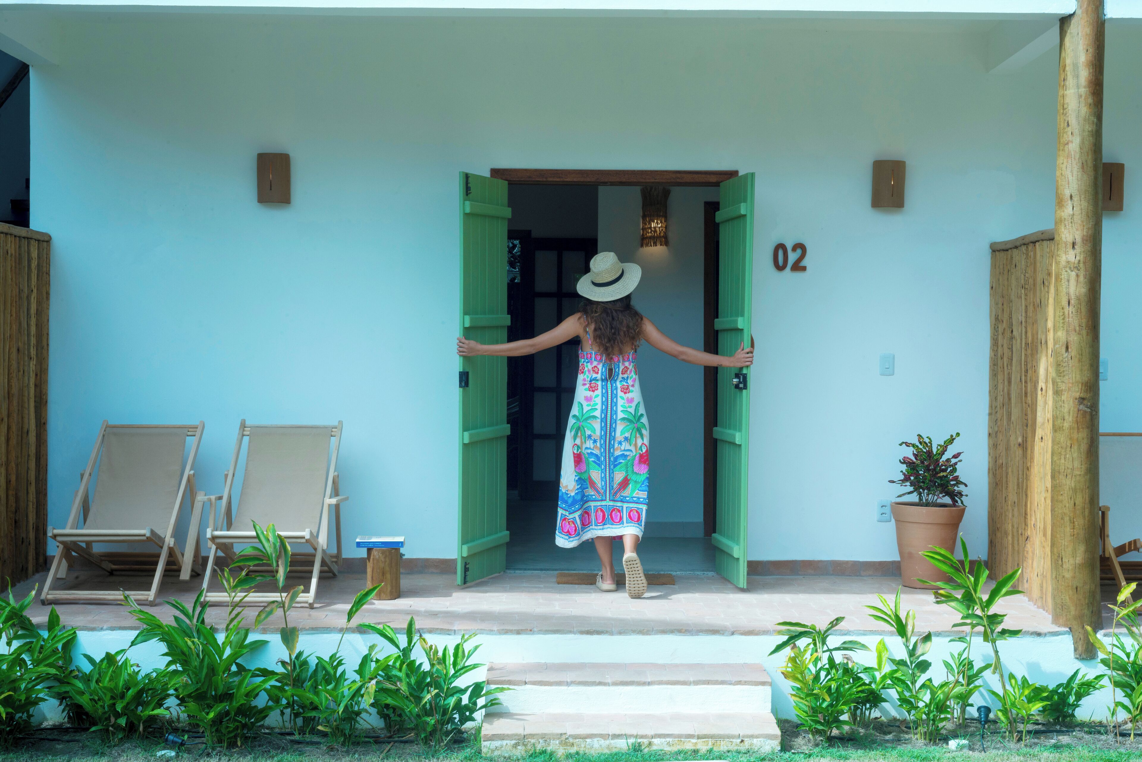 A woman in a colorful dress and hat entering a building through a green door. There are two wooden chairs and a potted plant outside.