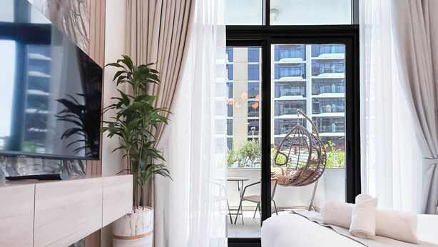 A modern hotel room with a large bed, a TV mounted on the wall, a potted plant, and a view of the city through the balcony doors. - Elegant Studio With Balcony on the Riviera Dubai (Dubai)