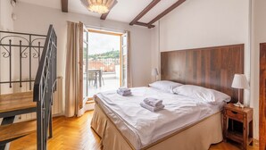 Un dormitorio con cabecera de madera, cama con sábanas blancas, mesita de noche con lámpara y vista a la ciudad a través de una puerta abierta.