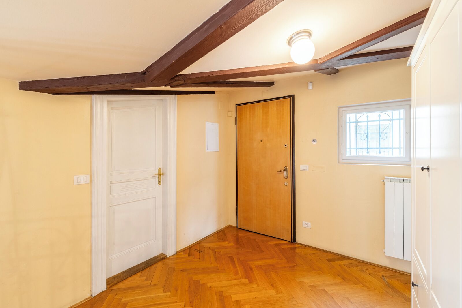 A hallway with wooden flooring, a white door, a wooden door, and a window with bars.