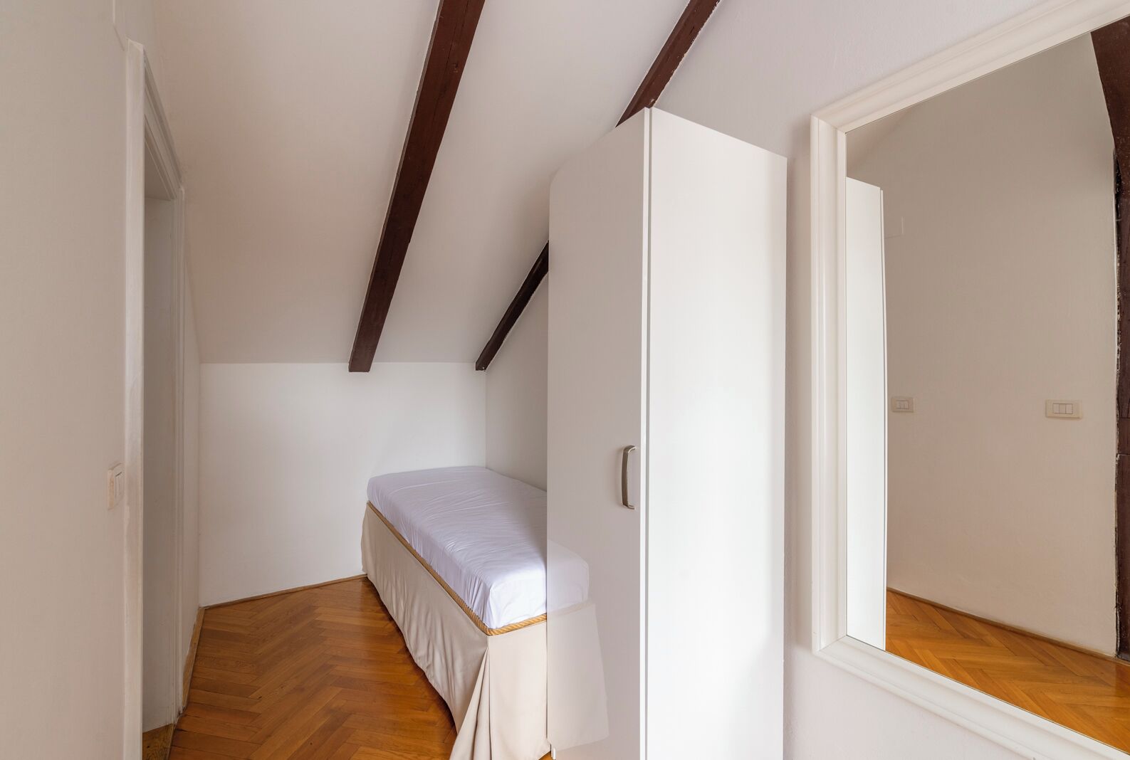 Una habitación pequeña con cama individual, suelo de madera, paredes blancas y techo inclinado con vigas a la vista.