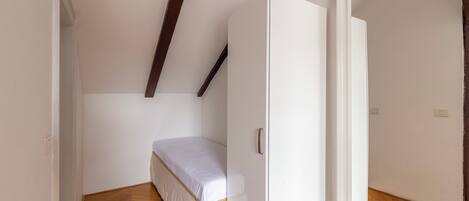 Una habitación pequeña con cama individual, suelo de madera, paredes blancas y techo inclinado con vigas a la vista.