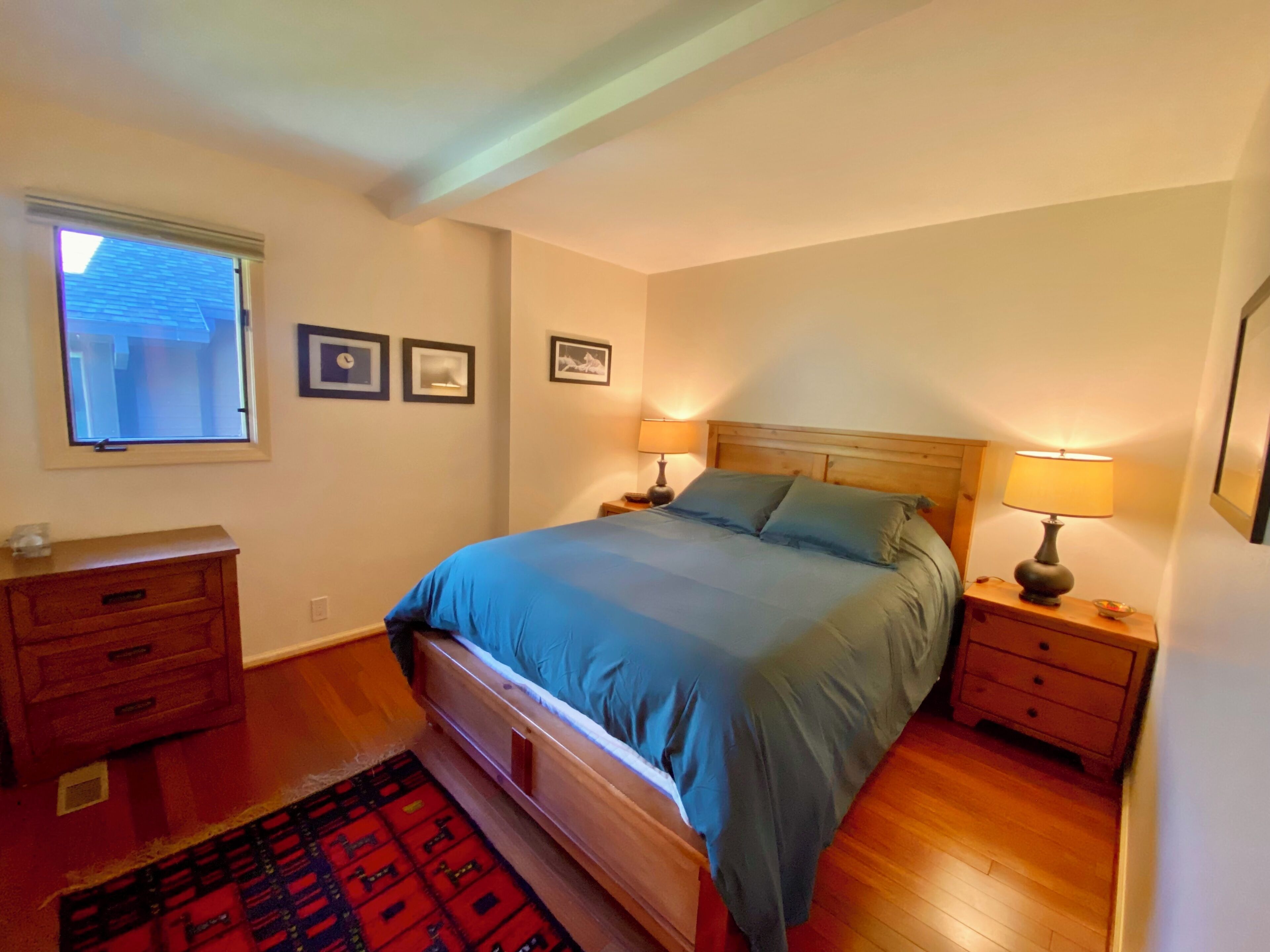 Une chambre à coucher comprenant un lit, deux tables de chevet avec des lampes, une commode, une fenêtre et trois tableaux encadrés accrochés au mur.