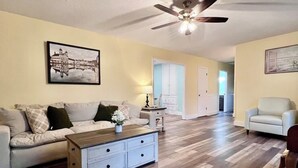 Un salon spacieux avec un plafond haut, un ventilateur de plafond et un plancher en bois franc. On y trouve un ensemble de canapés, une table basse et une porte qui donne sur une autre pièce.