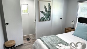Una camera da letto con un letto, un comodino, una lampada, una porta, un piccolo sgabello e una pianta in cornice.