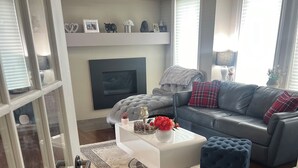 Een moderne woonkamer met een grijze bank, een witte salontafel en een blauwe poef. Er is een haard met daarboven een televisie, en een groot raam met luxaflex.