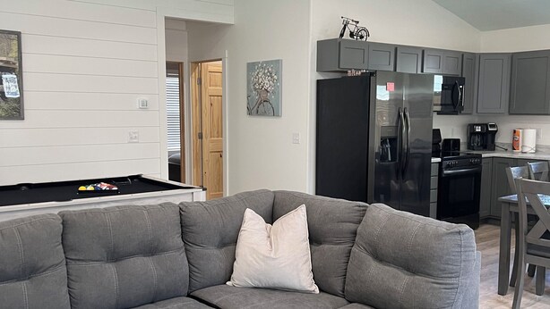 Una sala de estar moderna con un sofá seccional, un ventilador de techo y una cocina al fondo.