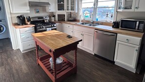 A modern kitchen with white cabinets, a wooden table, stainless steel appliances, and a view of the outside through a window. - Cozy cottage in Hatteras Village. Convenient to shopping, restaurants and beach. (Hatteras)
