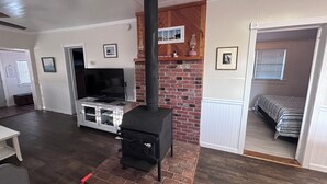 A living room with a fireplace, a television, and a bed in a separate room. - Cozy cottage in Hatteras Village. Convenient to shopping, restaurants and beach. (Hatteras)