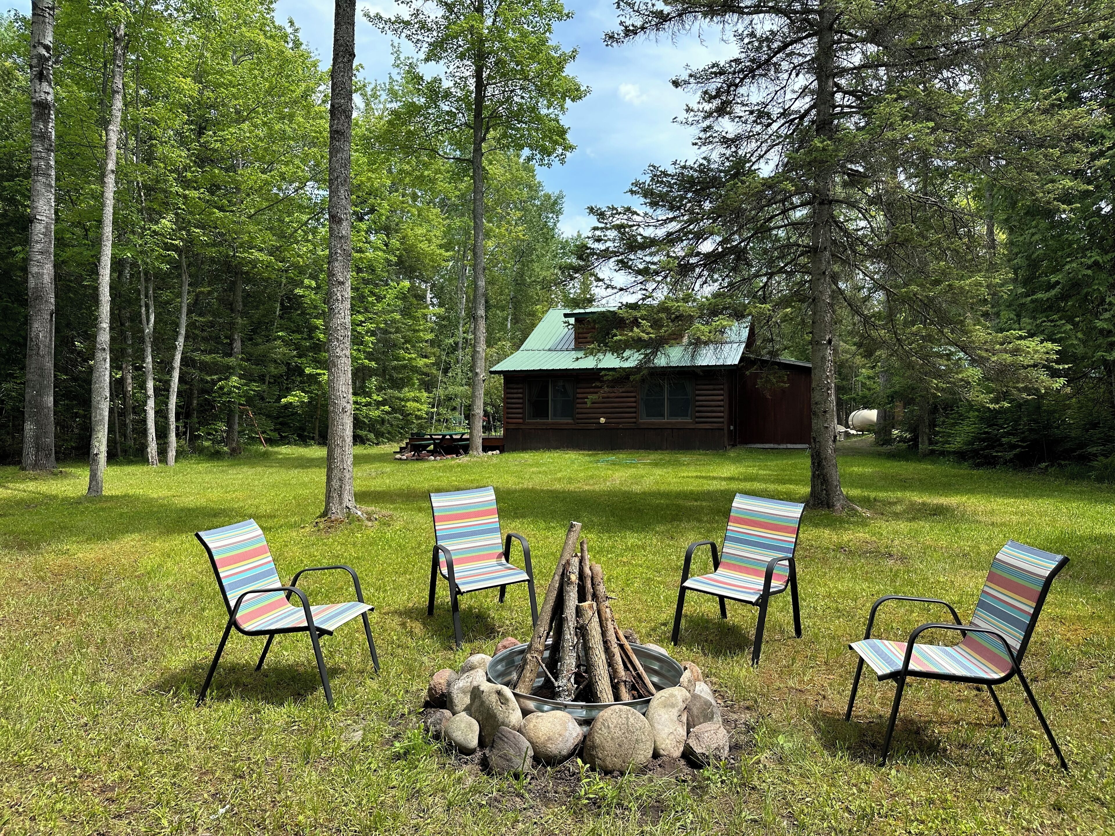 Une cabane entourée d’une zone herbeuse, avec quatre chaises à rayures disposées autour d’un foyer.