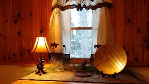 A wooden interior with a table, a lamp, a decorative plate, and a window with a view of trees.
