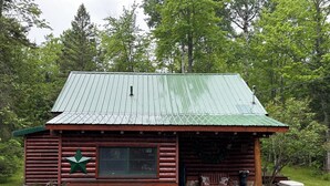 A rustic log cabin with a green metal roof, a stone foundation, and a porch with a bench.