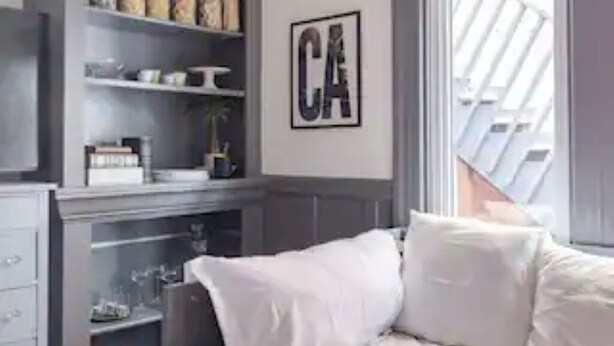 A bedroom with a bed, a grey wardrobe, a shelf with jars, and a framed letter 'CA' on the wall.