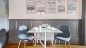 A dining area with a white table and two blue chairs, framed pictures on the wall, and wooden flooring.