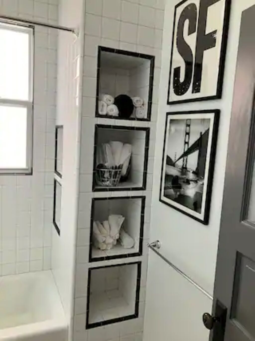 A bathroom with white tiled walls, a bathtub, and a shelf unit with various items.