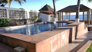 A rectangular swimming pool with a stone border, a thatched-roof structure, and a wooden gazebo by the sea.