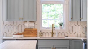 Une cuisine avec des meubles de couleur bleu clair, un plan de travail blanc, une planche à découper en bois, un robinet doré et une fenêtre donnant sur un paysage verdoyant.