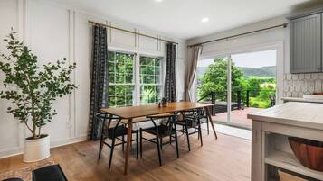A dining area with a wooden table and chairs, a potted plant, and a view of greenery through large windows.