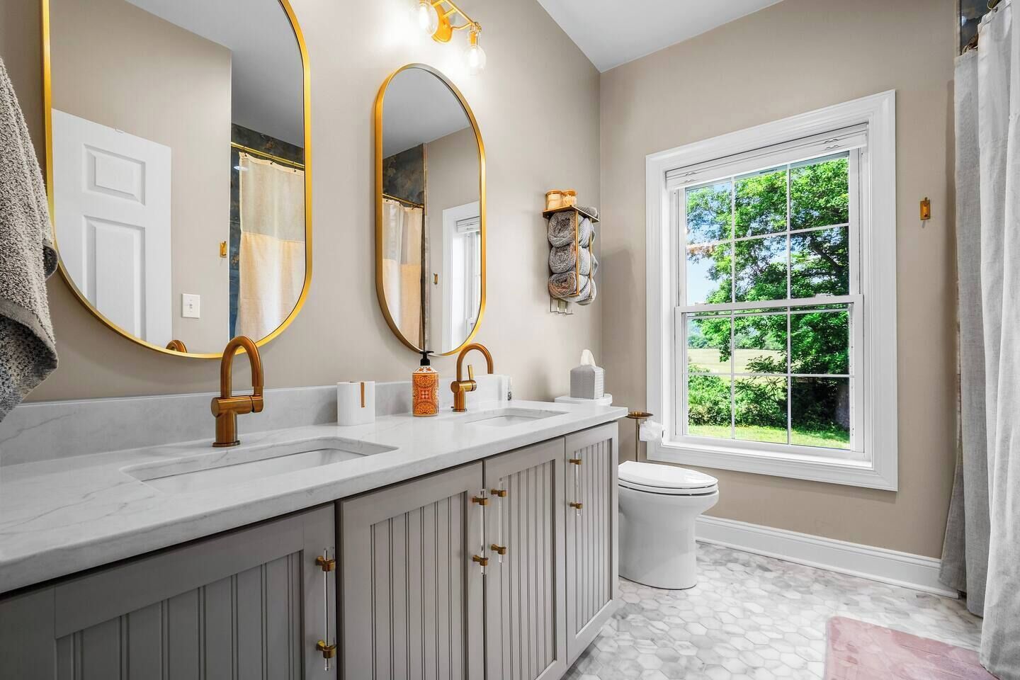A bathroom with a double sink vanity, a toilet, and a window with a view of greenery.