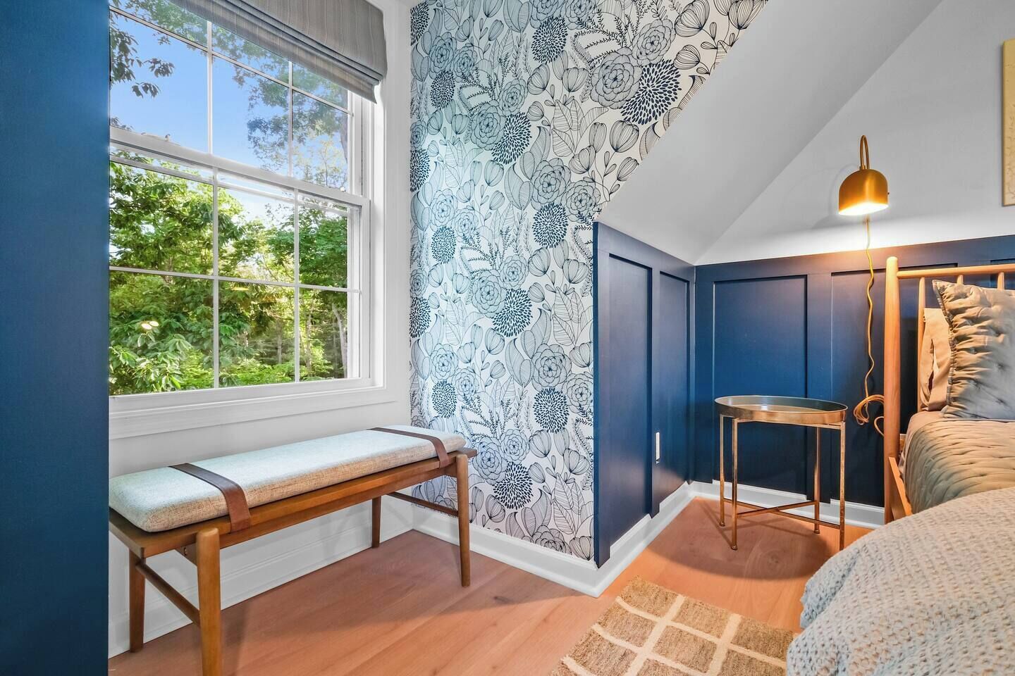 A bedroom with a bench, a bedside table, a lamp, and a view of greenery through a window.