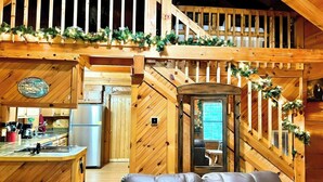 A wooden cabin interior with a kitchen, dining area, and a staircase leading to a loft. - Cozy Cabin w/ 2 King Beds, Fireplace, Hot Tub, Foosball, Arcade and BBQ! (Sevierville)