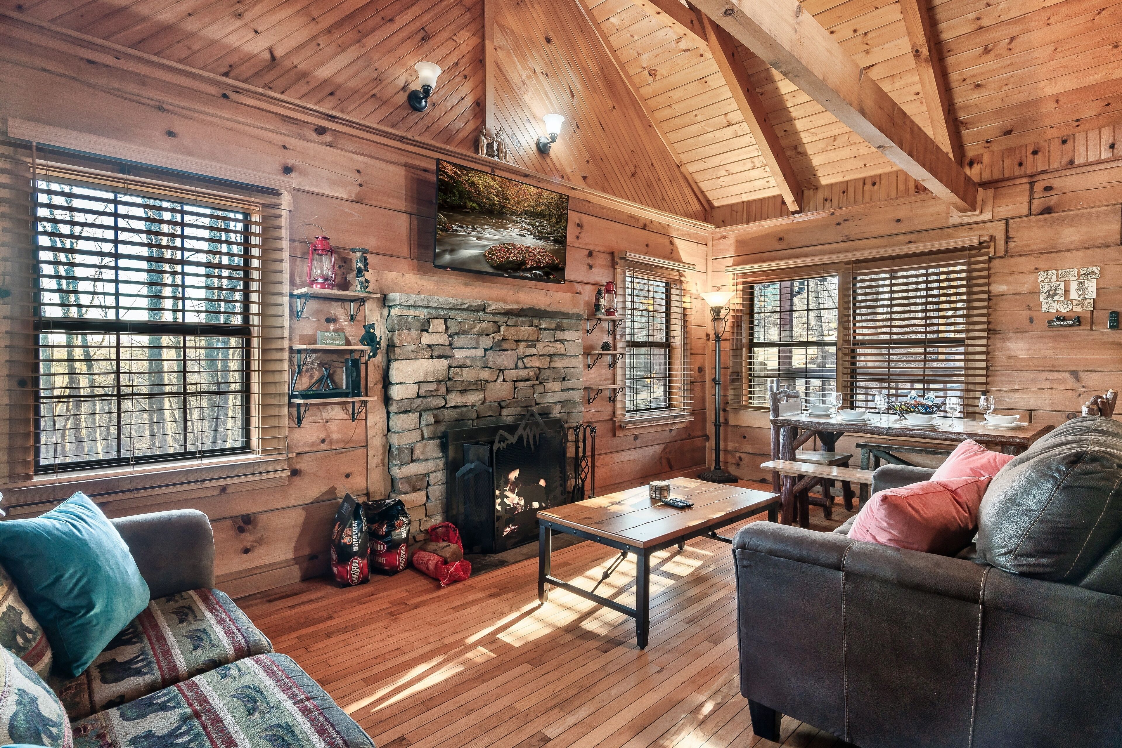 A cozy log cabin interior with a stone fireplace, wooden furniture, and large windows.