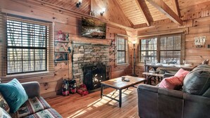 A cozy log cabin interior with a stone fireplace, wooden furniture, and large windows. - Cozy Cabin w/ 2 King Beds, Fireplace, Hot Tub, Foosball, Arcade and BBQ! (Sevierville)