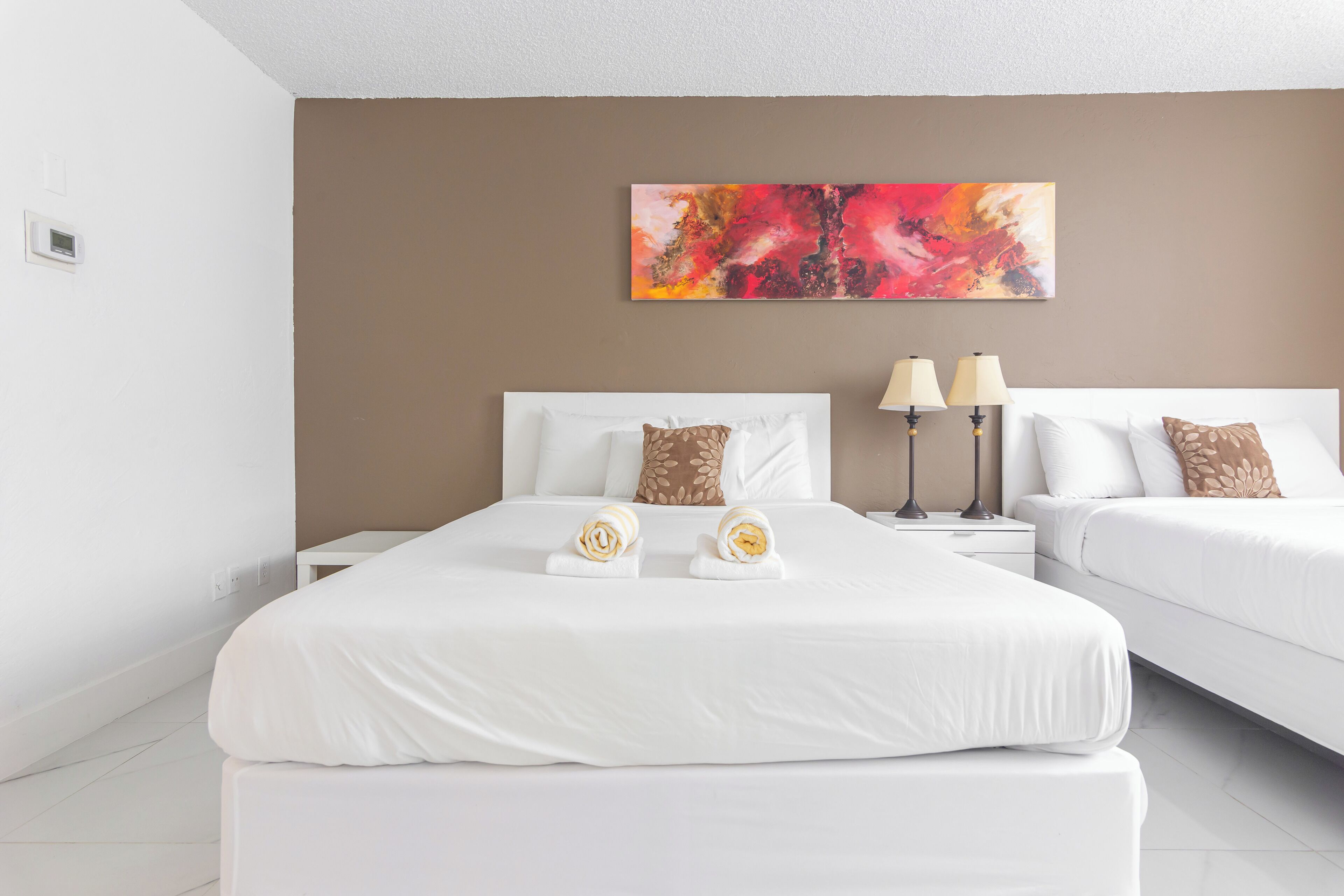 Quarto de hotel moderno com duas camas, uma pintura abstrata grande e uma mesa de cabeceira com duas luminárias.