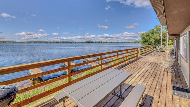 Una terraza de madera con barandilla que da a un lago, una mesa de picnic y un área cubierta con sillas.