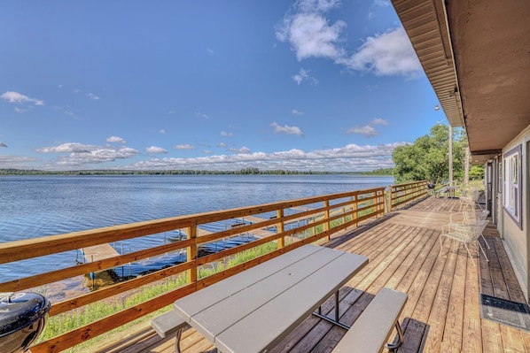 A wooden deck with a railing overlooking a lake, a picnic table, and a covered area with chairs.
