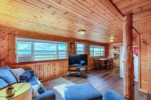 Eine Hütte mit Fernseher, Sitzecke, Esstisch und großen Fenstern mit Blick auf das Wasser.