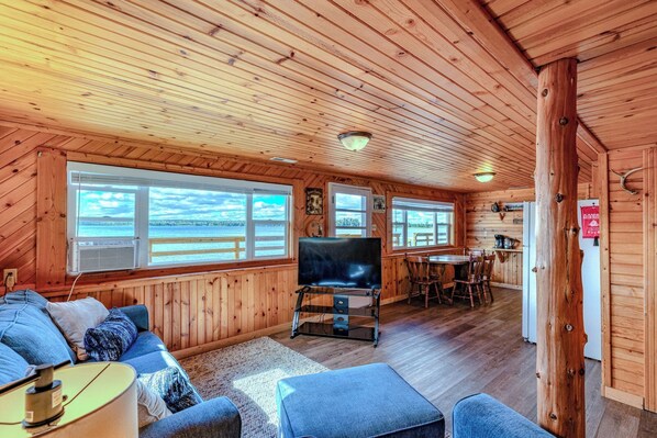 A cabin with a TV, seating area, dining table, and large windows overlooking water.