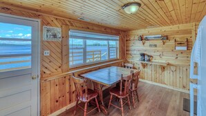 Interior de una cabaña de madera con una mesa de comedor, sillas, ventana con vista al agua y techo de madera.
