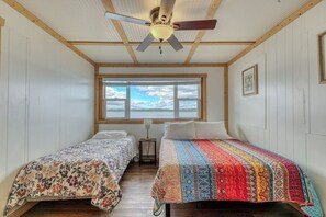 A room with two single beds, a ceiling fan, a window with a view, and a framed picture on the wall.