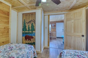 A room with two beds, a wooden ceiling, a door leading to a kitchen, and a wall with a vertical textile artwork. - Cabin on the Lake - 4-Beds - Open for Hunting/Rifle Season - Pets Ok (Gordon)