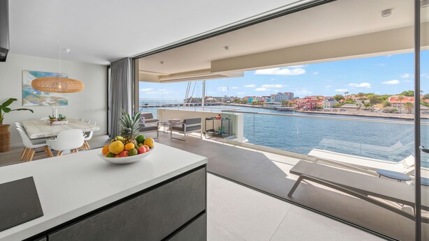 Et moderne køkken med skålsæt med frugt, spiseplads og en balkon med udsigt over vandet og bybilledet.