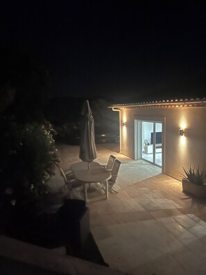 Un espace patio avec une table et des chaises, un parasol et un petit bâtiment doté d’un éclairage extérieur.