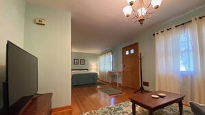 A living room with a television, a wooden coffee table, a sofa, and a dining table. There is a bed in the background and a door leading to another room. - Clintonville Charmer | Walk to Great Restaurants! (Columbus)