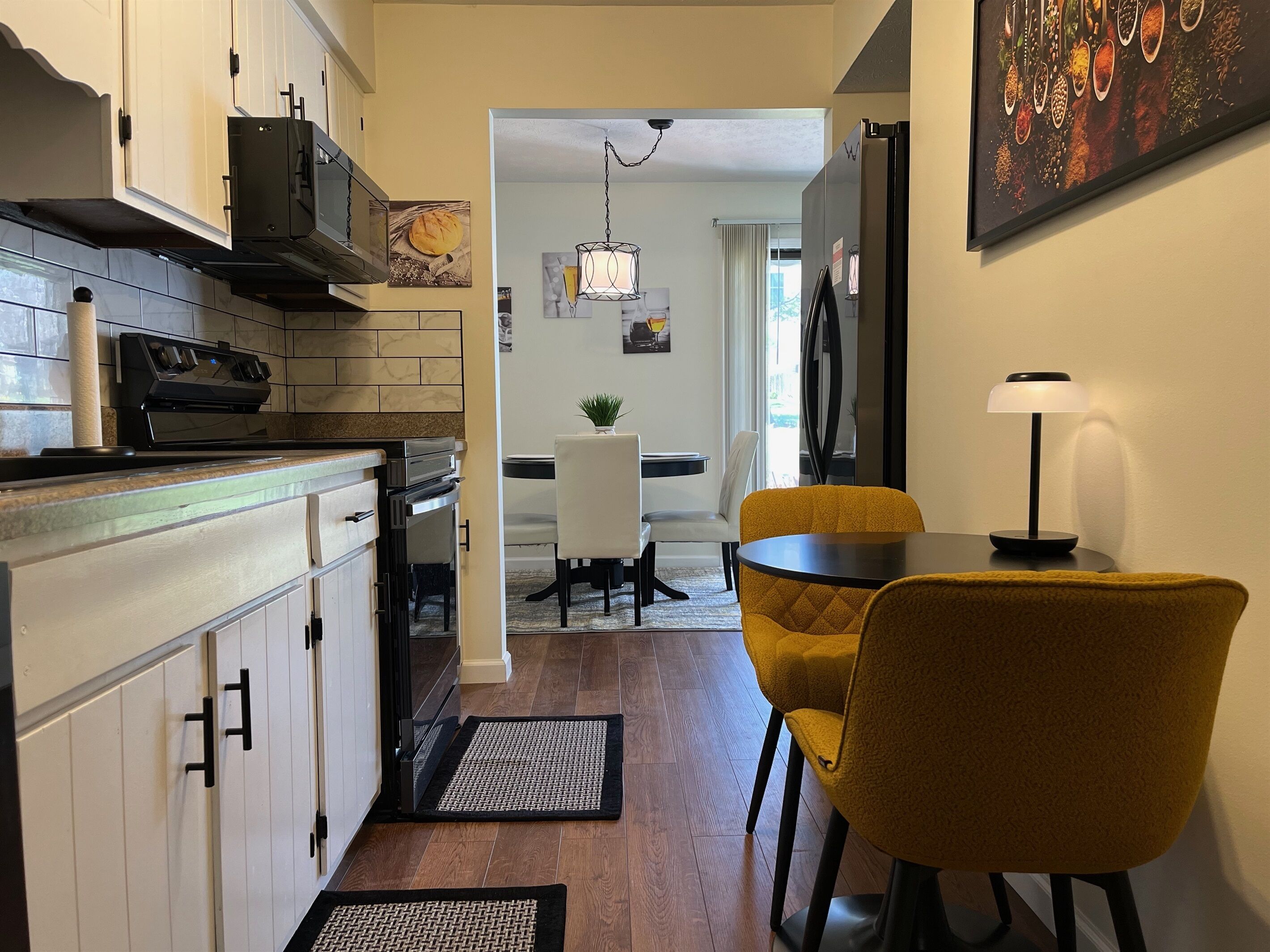 A modern kitchen with white cabinets, a black range hood, and a tiled backsplash. There is a dining area with a yellow chair and a table, a small plant on the table, and a painting on the wall.