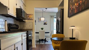 A modern kitchen with white cabinets, a black range hood, and a tiled backsplash. There is a dining area with a yellow chair and a table, a small plant on the table, and a painting on the wall. - Westerville Comfort & Convenience (Westerville)