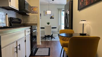 A modern kitchen with white cabinets, a black range hood, and a tiled backsplash. There is a dining area with a yellow chair and a table, a small plant on the table, and a painting on the wall.