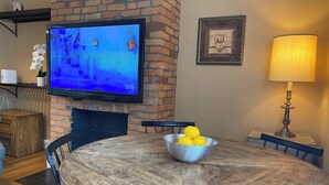 En matplats med ett runt träbord, en skål med citroner, en tv som är monterad på en tegelvägg och en lampa till höger.
