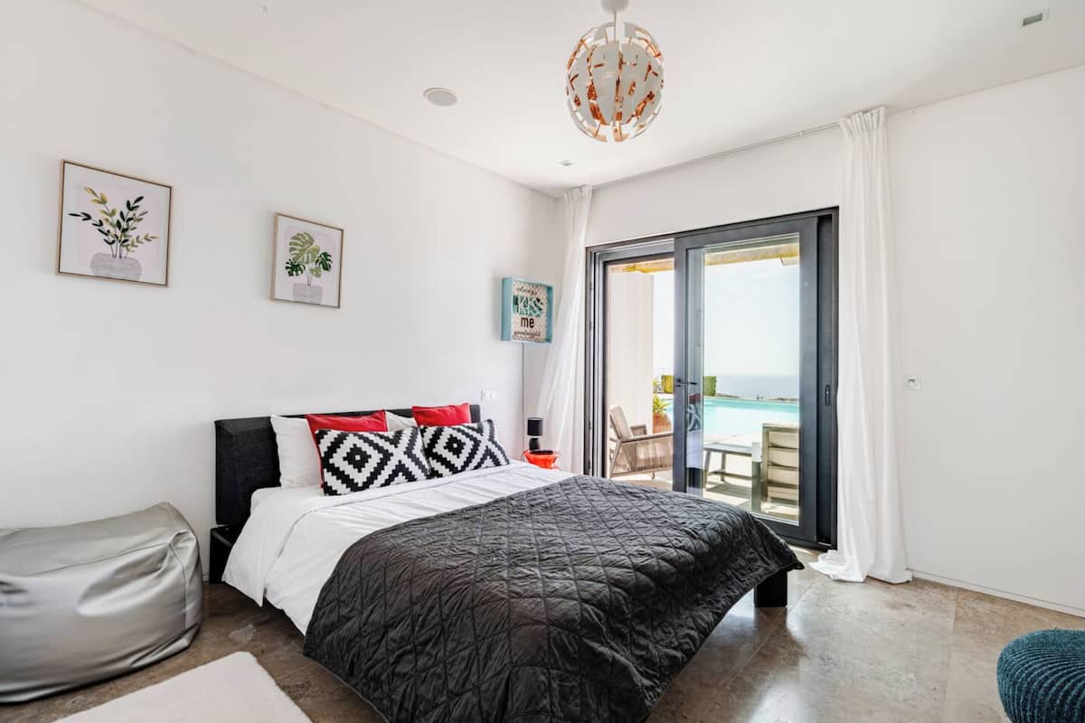 Une chambre avec un lit, une table de chevet, une chaise, un fauteuil poire et une vue sur la mer, visible à travers une porte-fenêtre coulissante.