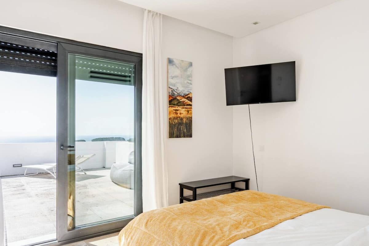 Une chambre dotée d’un grand lit, d’un téléviseur fixé au mur et d’une porte-fenêtre coulissante donnant sur un espace extérieur avec une chaise blanche.