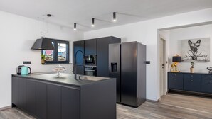 Une cuisine moderne dotée d’un îlot central, de meubles de rangement noirs, d’un réfrigérateur en acier inoxydable et d’un luminaire suspendu.