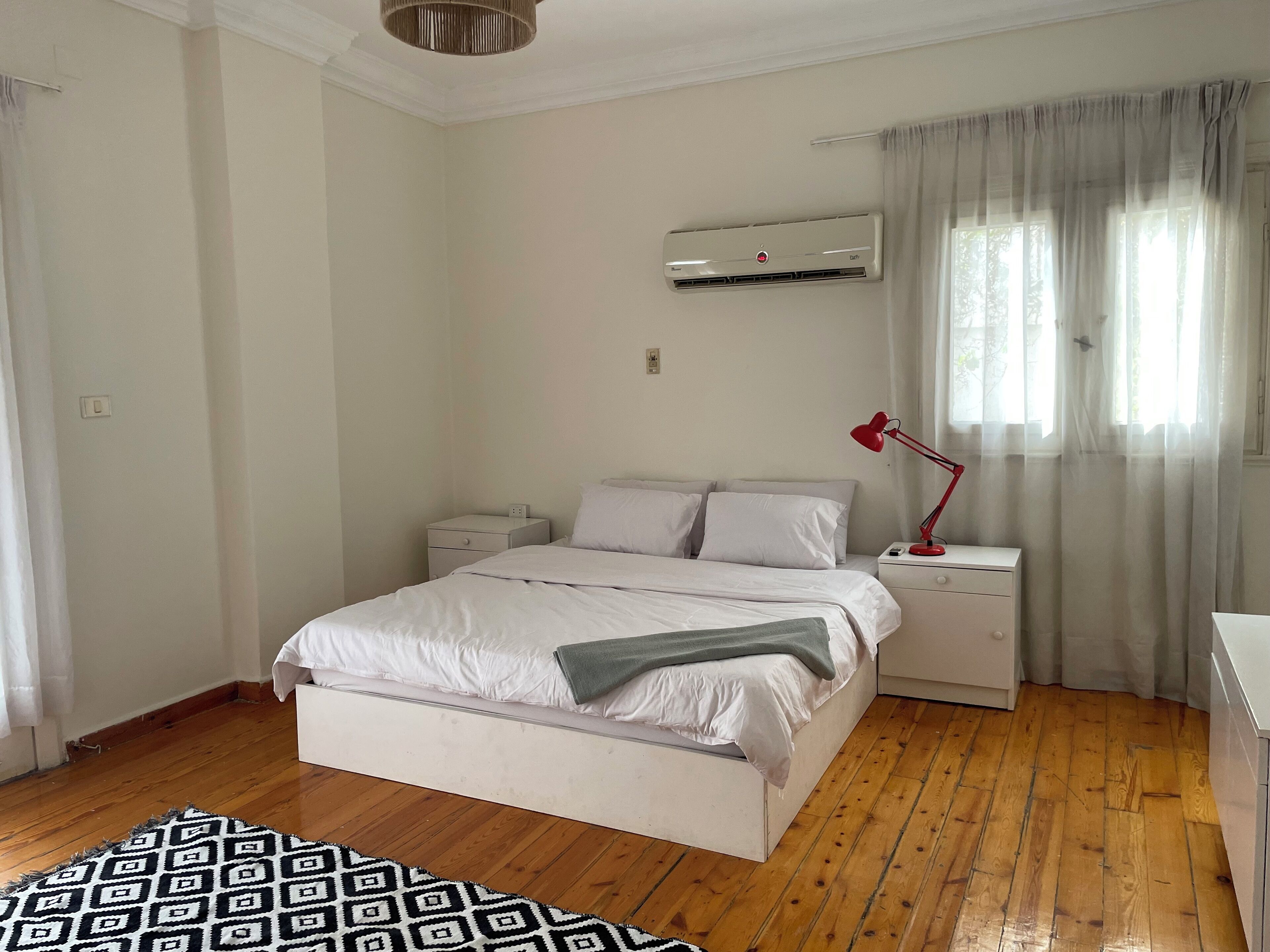 A bedroom with a bed, bedside tables, a red lamp, an air conditioner, and a patterned rug.