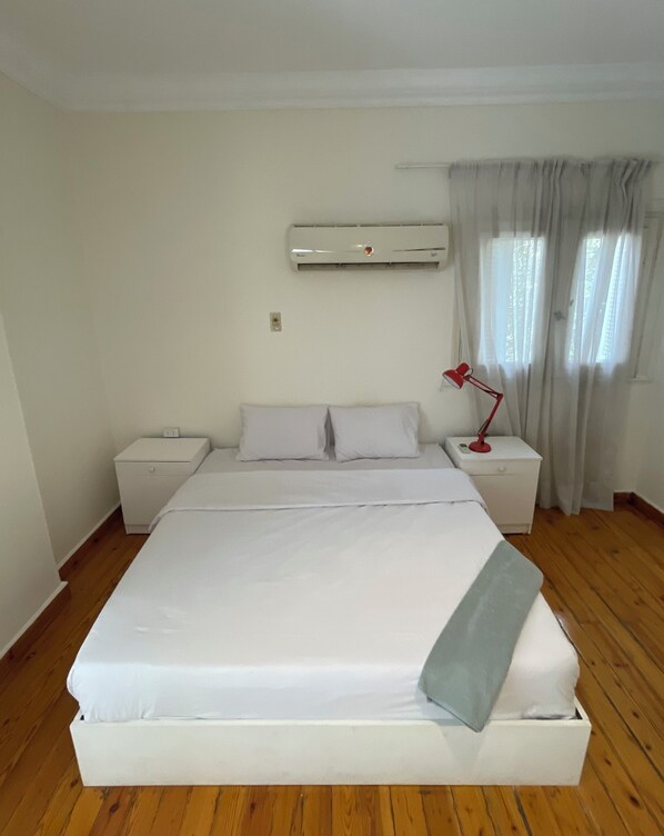 A bedroom with a large bed, two bedside tables, a red desk lamp, an air conditioning unit, and a window with curtains.