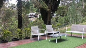Un patio con mobiliario exterior blanco, césped artificial verde y una terraza de madera.