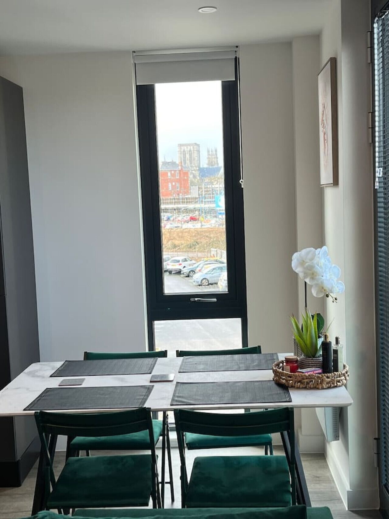 Un comedor con una mesa dispuesta para cuatro, una ventana que ofrece vistas a edificios y automóviles estacionados, y una planta decorativa en el alféizar.